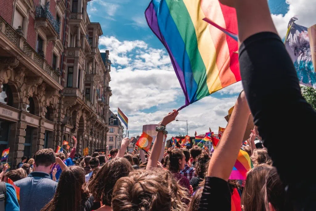 A Pride parade in 2023 marching the streets of Strasbourg, France waving rainbow flags against a blue cloudy sky.