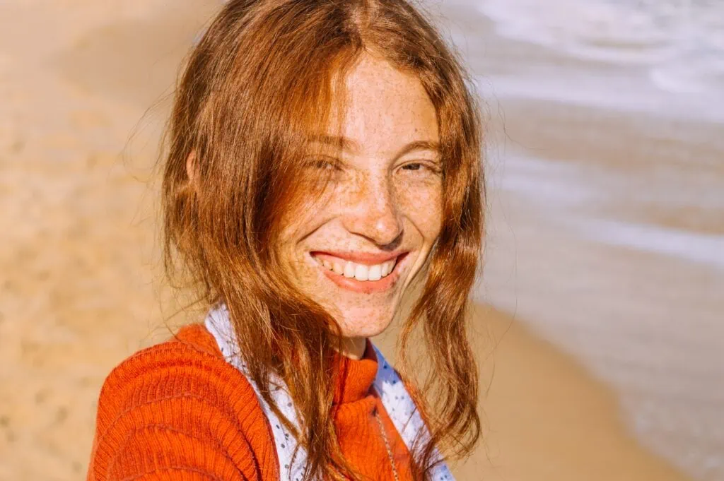A close-up of a freckled woman at the beach smiling at the camera.