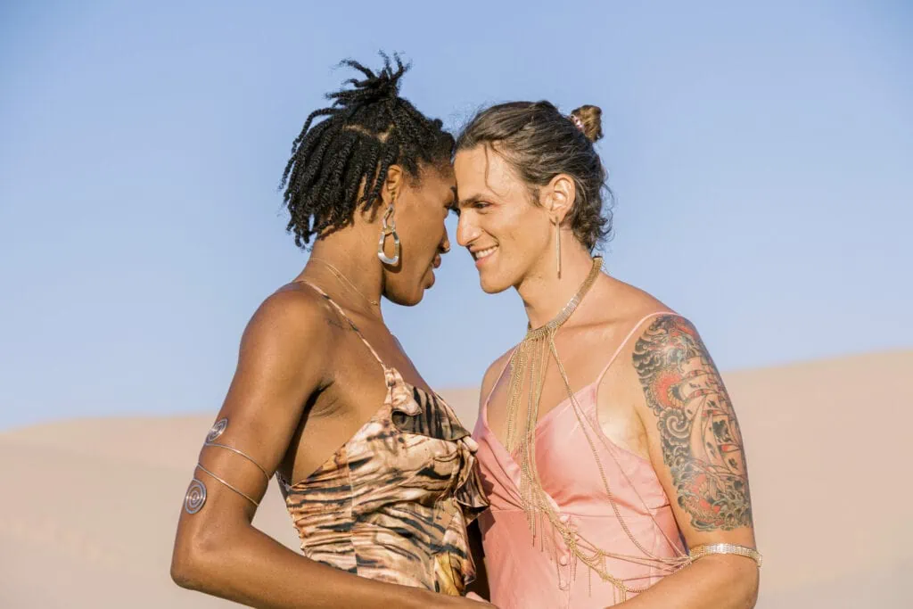 A couple of women in pretty dresses staring at each other lovingly, the blurred background looks like a desert.