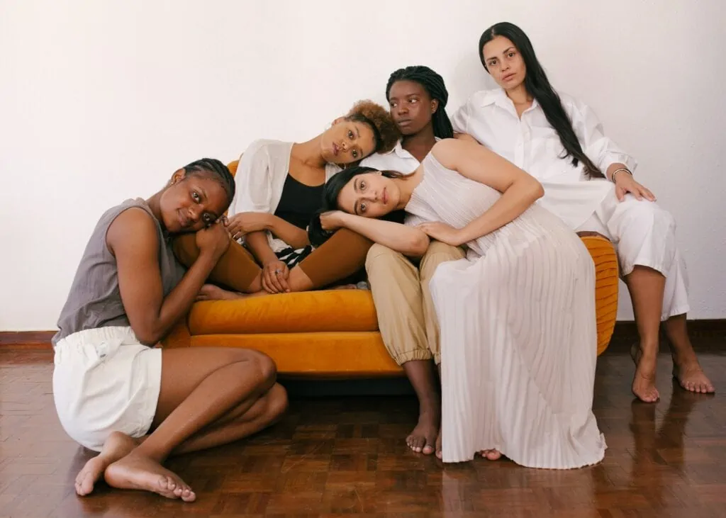 A diverse group of women, 3 black and 2 indigenous, sit on a couch holding onto one another