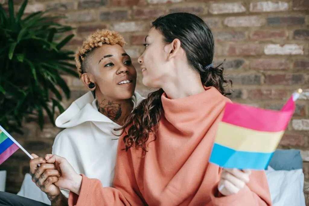 A black woman and a white woman look into each other's eyes. The white woman holds a pansexual flag