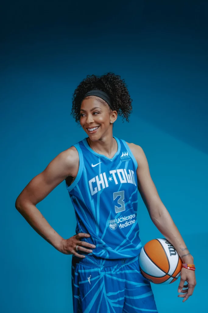 Candace Parker against a blue backdrop wearing a blue basketball uniform, smiling, and holding a basketball against her left arm.