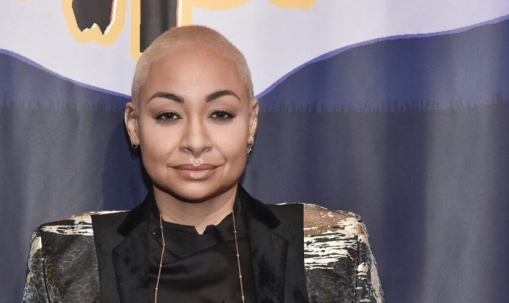 Raven-Symoné wearing a cool patterned tuxedo on a red carpet, smiling gently and looking radiant.