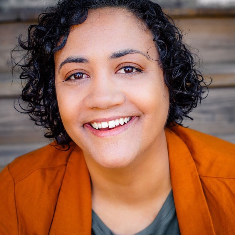 Allison Reese is a lesbian comedian of color who has guest starred in the movie Bros and recently headlined at Do Better Comedy in Pheonix, Arizona, seen in this image. This picture shows Allison on stage facing the crowd at Do Better, performing in front of a decorated Christmas tree. She wears a baseball cap with a graphic t-shirt and a striped button-down on top, while holding and speaking into the microphone.