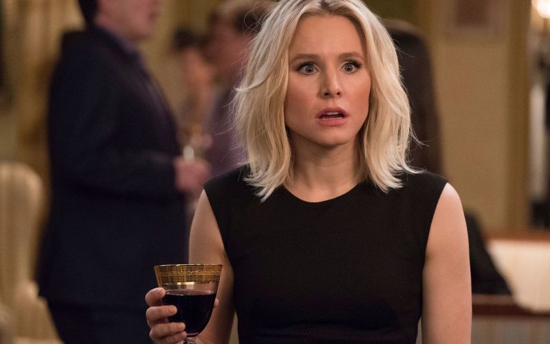 Eleanor is standing with a glass of wine in her hand, her mouth open in shock.