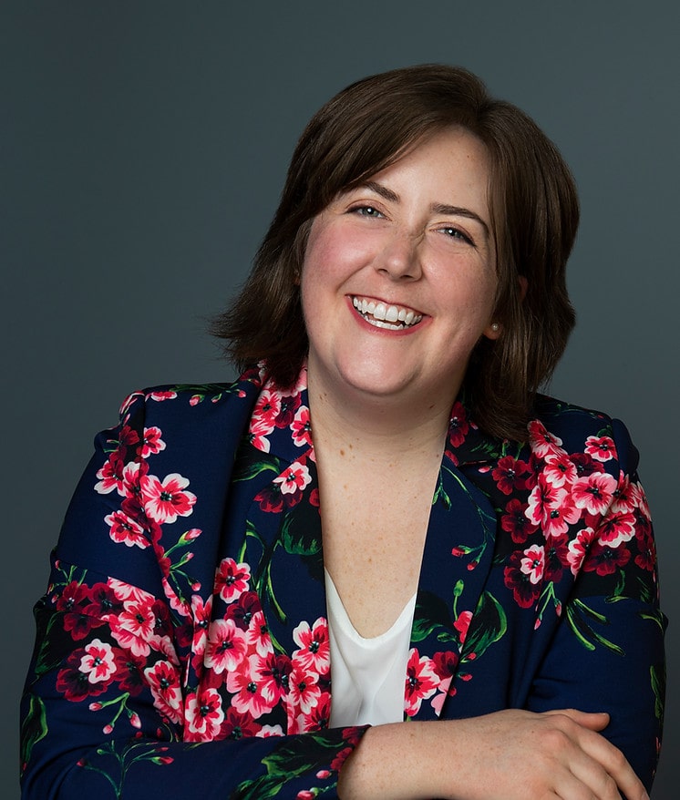 Katie Nixon is a gay female comedian who originally started her career in Chicago. She is now a cast member of BOOM Chicago, a comedy club in Amsterdam. She’s known for her musical comedy and is seen in this headshot smiling with her arms crossed. She wears a white top and a floral blazer while donning medium-length hair. 
