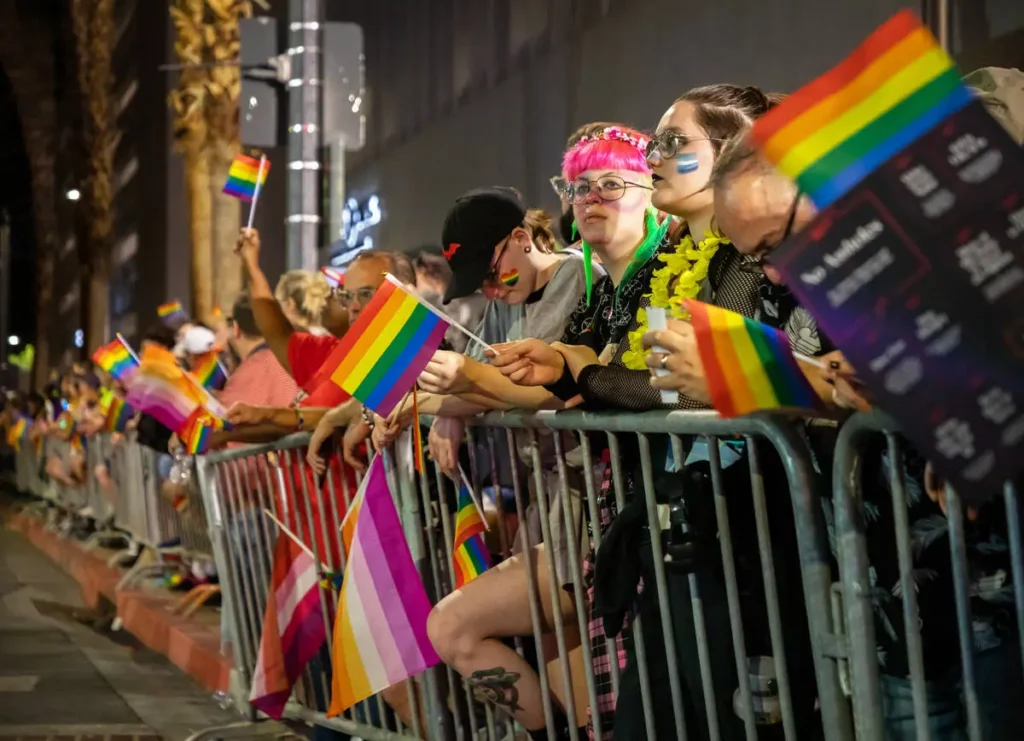 A crowd of people stand behind barriers. They are holding lesbian and gay pride flags and donning pride colored makeup at the Las Vegas PRIDE parade.