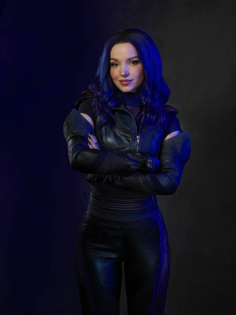 Mal stands with arms crossed facing the camera. Mal, played by Dove Cameron, is a villain-turned-hero from the Disney franchise Descendants. She’s characterized by having long dark purple hair, normally black or purple clothing.