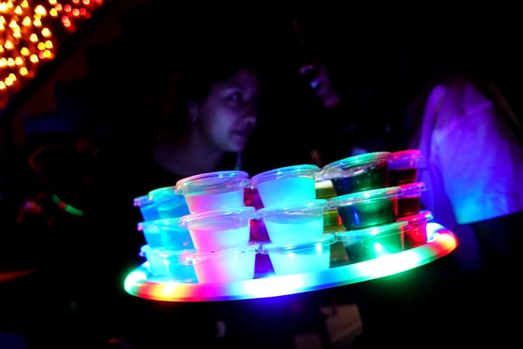 Get your jello shots at Number One Fifth Avenue, a gayborhood staple in San Diego. Dancing, music, parties, and a mixed crowd on some nights, are all that you can expect. Here you see stacks of jello shots on a circular tray with neon lighting, making the jello look fluorescent. Two darkly-lit women stand behind the tray, speaking to each other, at Number One Fifth Avenue.