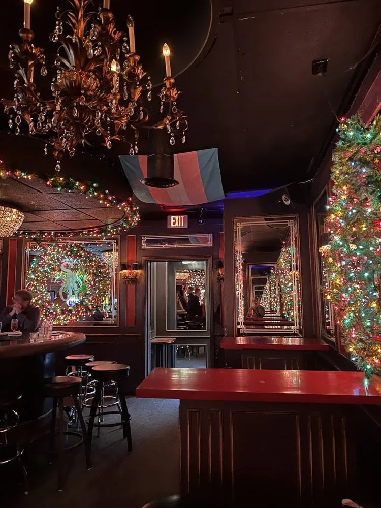 An interior view of the SRO Lounge when it is mostly empty. It has ornate decorations, like a fake candle chandelier, and Christmas lights. A transgender pride flag is hanging right above the bar’s exit. The SRO Lounge is a place for lesbians and trans people to meet and feel safe.