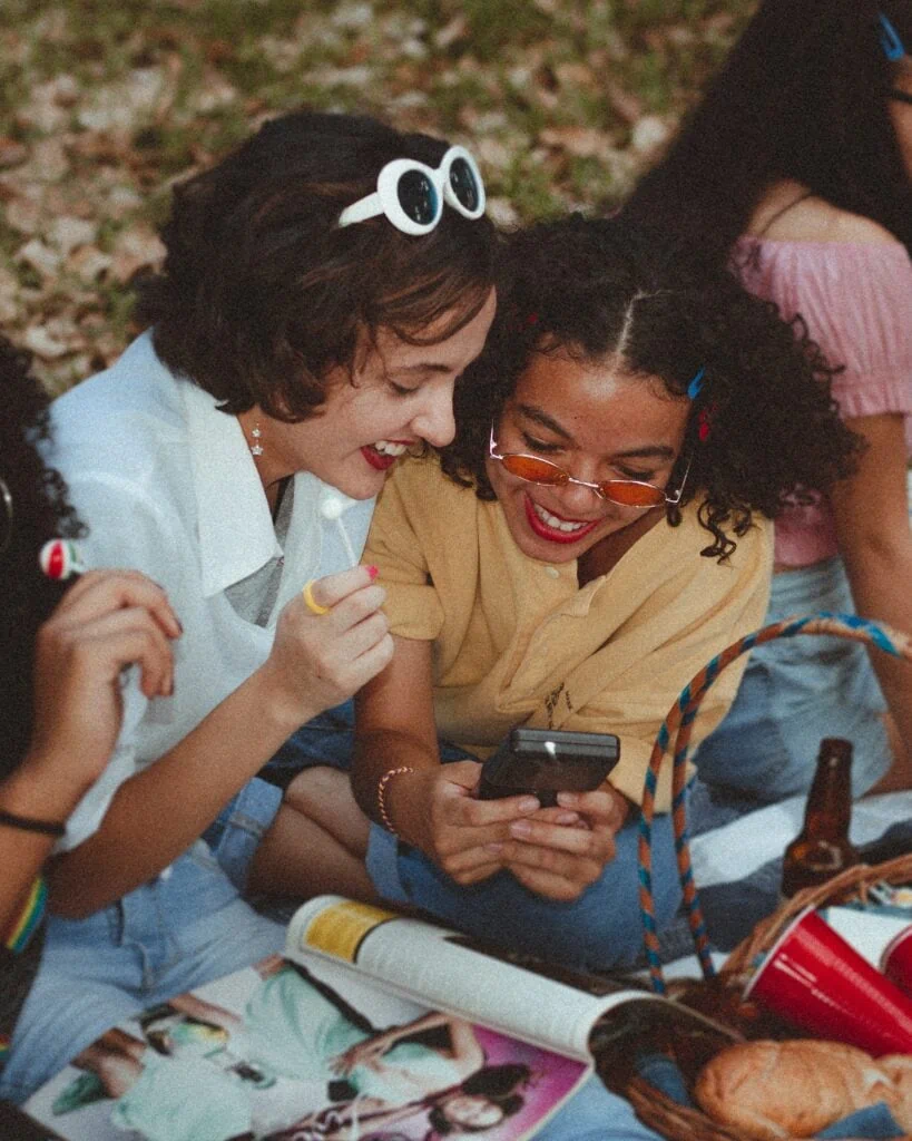 The image is a shallow focus photograph of a woman using a Game Boy, smiling next to another woman. They are on a picnic blanket surrounded by a picnic basket with bread and red solo cups. There are also beer bottles and magazines and other women around them. The San Diego Meetup groups hosts picnics and game themed events like in the photo.