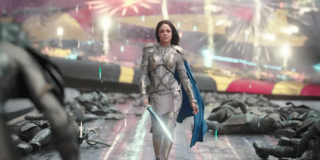 Valkyrie, played by queer actress Tessa Thompson, walks toward the camera confidently in front of a battle and fallen soldiers.
