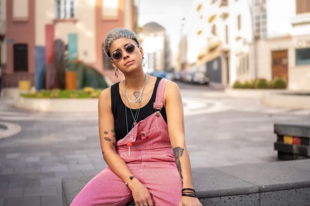 A stylish young queer person sitting on a ledge with sunglasses and overalls.