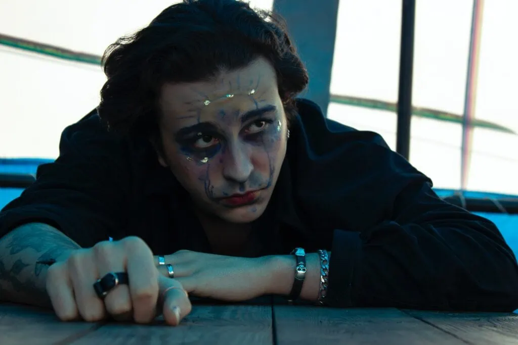 A gender-fluid person with jewelry and makeup drawn on their face, while laying down.