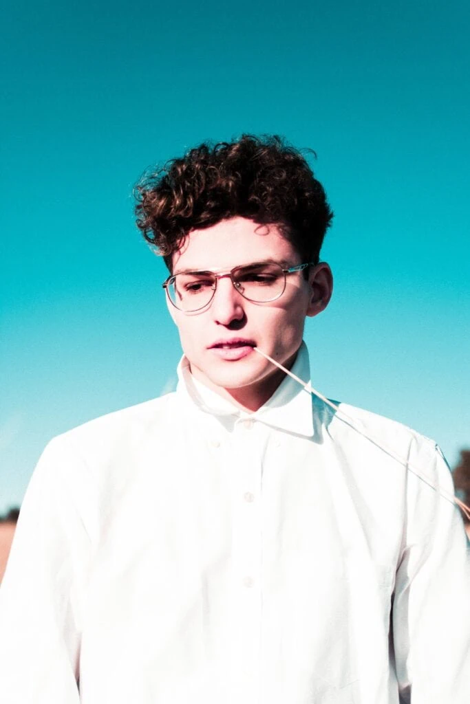 An androgynous person standing in front of a blue sky with a white shirt on.