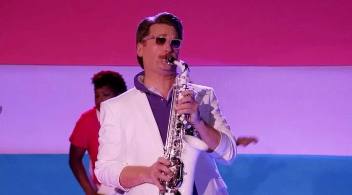 Pete Gardner plays the saxophone, wearing sunglasses in front of the bisexual flag.