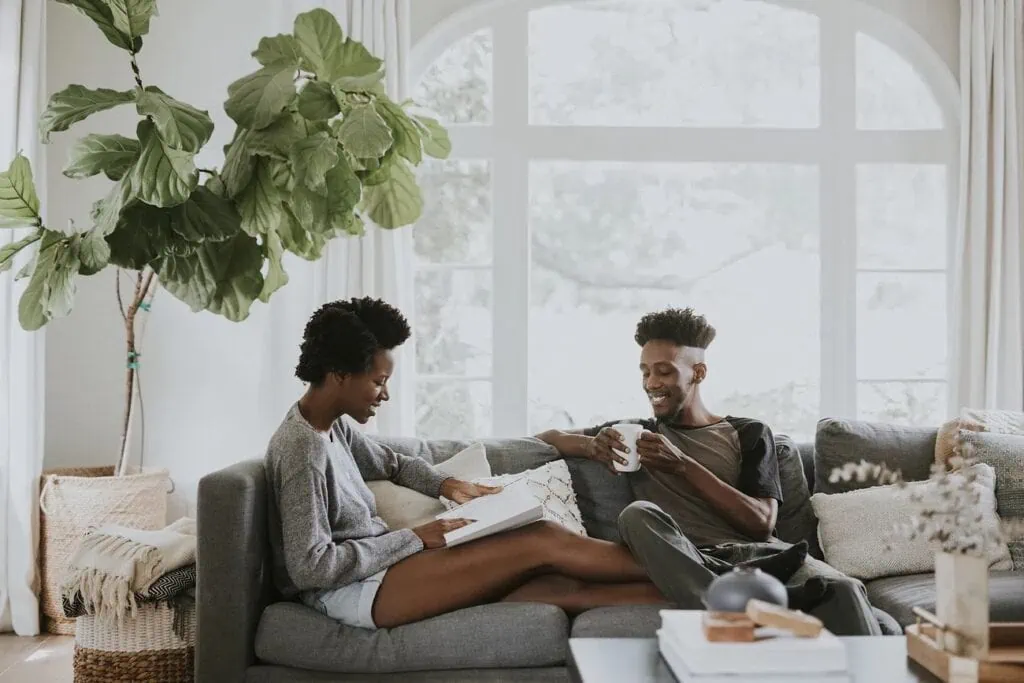 A couple sit at-ease on a couch in their home. One of them holds a cup and smiles at the other person who reads a book out loud. 