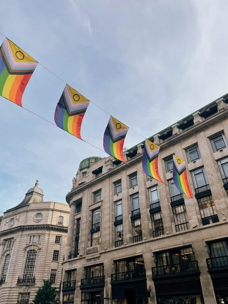 Intersex-inclusive progress pride flags hang between buildings in a city. This flag has the six rainbow stripes, with a chevron pattern of brown, black, pink, blue, and white stripes, as well a purple circle superimposed on a yellow triangle.
