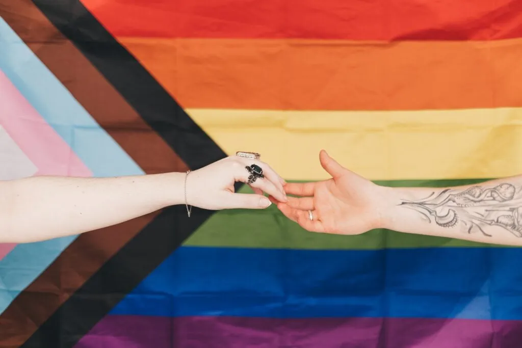 Two hands reach for each other in front of the pride progress flag, which has the six colors of the LGBT pride flag and a chevron on the left-hand side with black, brown, blue, pink, and white stripes. 
