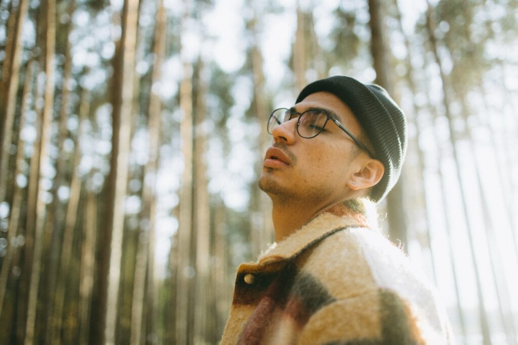 A person with glasses wears a black beanie. Tall trees take up the background of the photo.