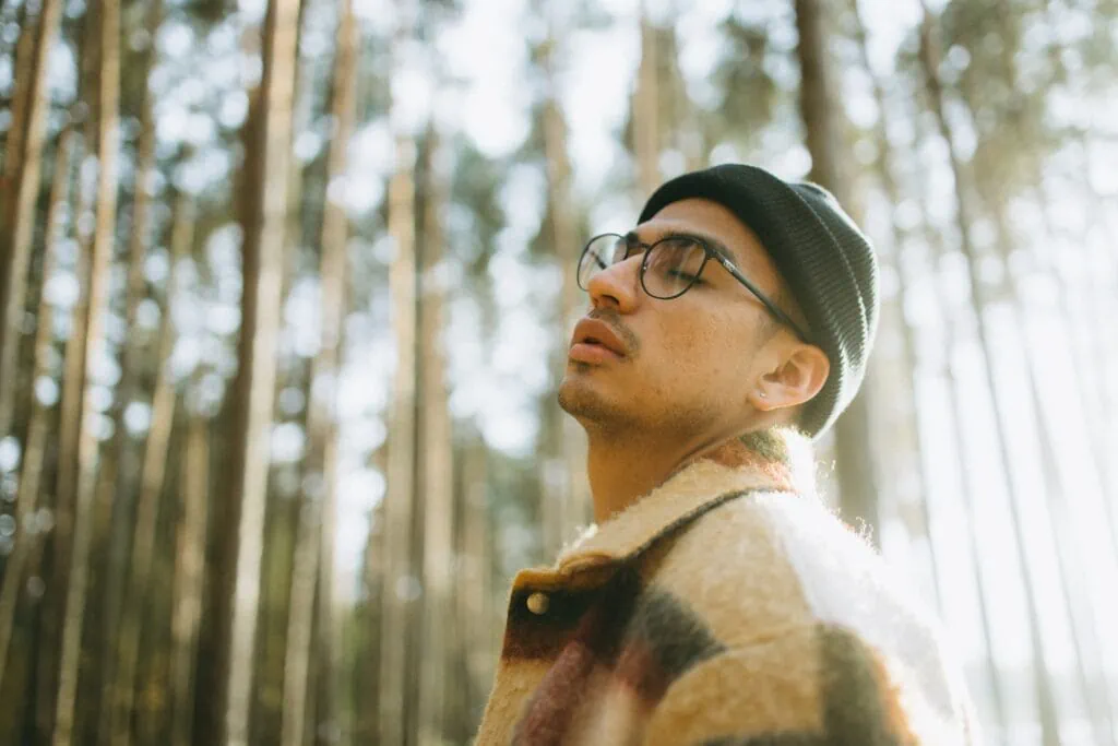 A person with glasses wears a black beanie. Tall trees take up the background of the photo.