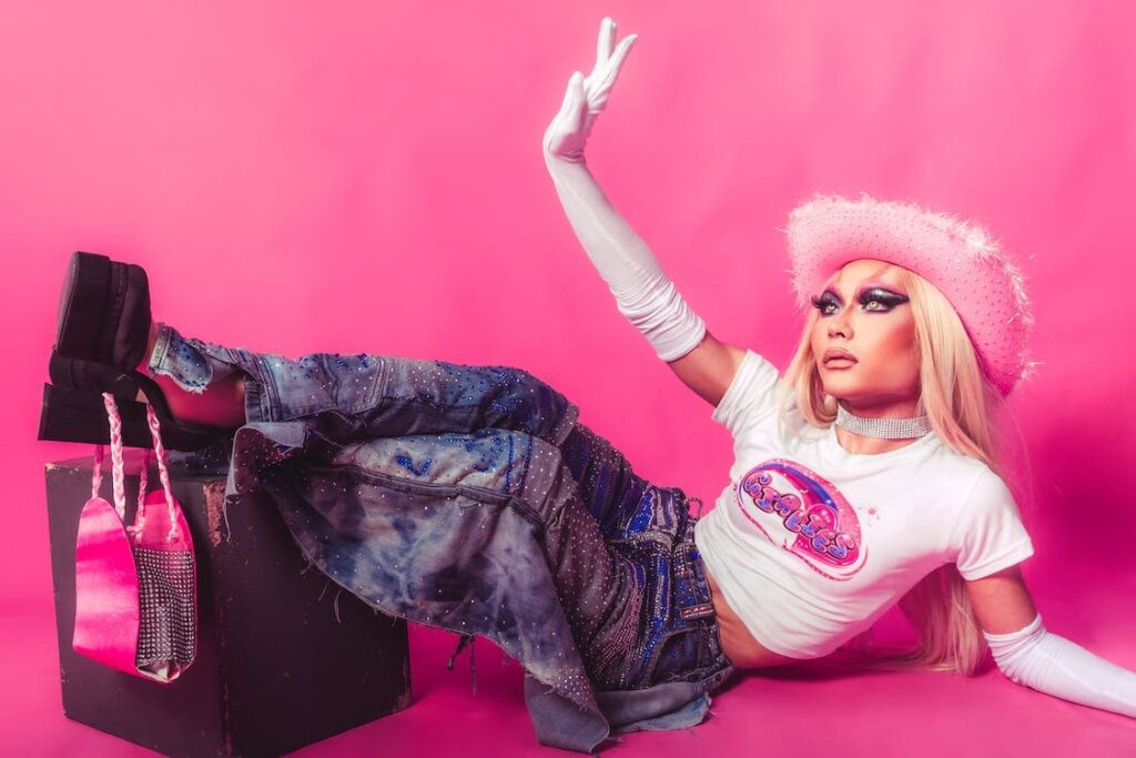 A person lays in front of a pink background. They wear a pink hat, crop top, and bedazzled distressed jeans and platform shoes