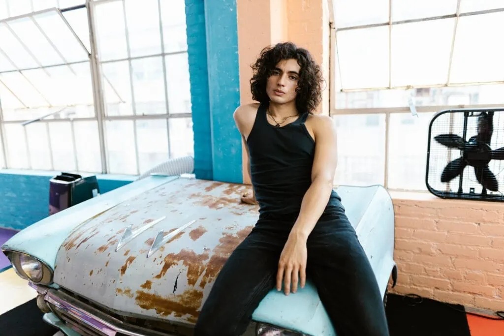 A stylish person with a curly bisexual bob sits on the hood of a car.
