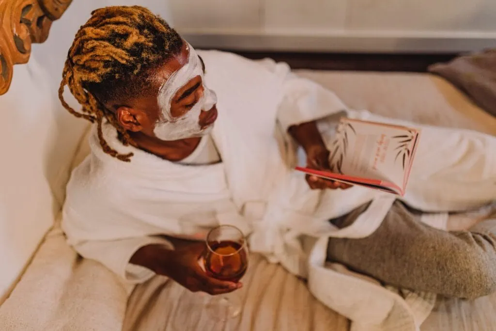A person lays on a bed with rosé and a book in hand. They have a face mask and a bath robe on.