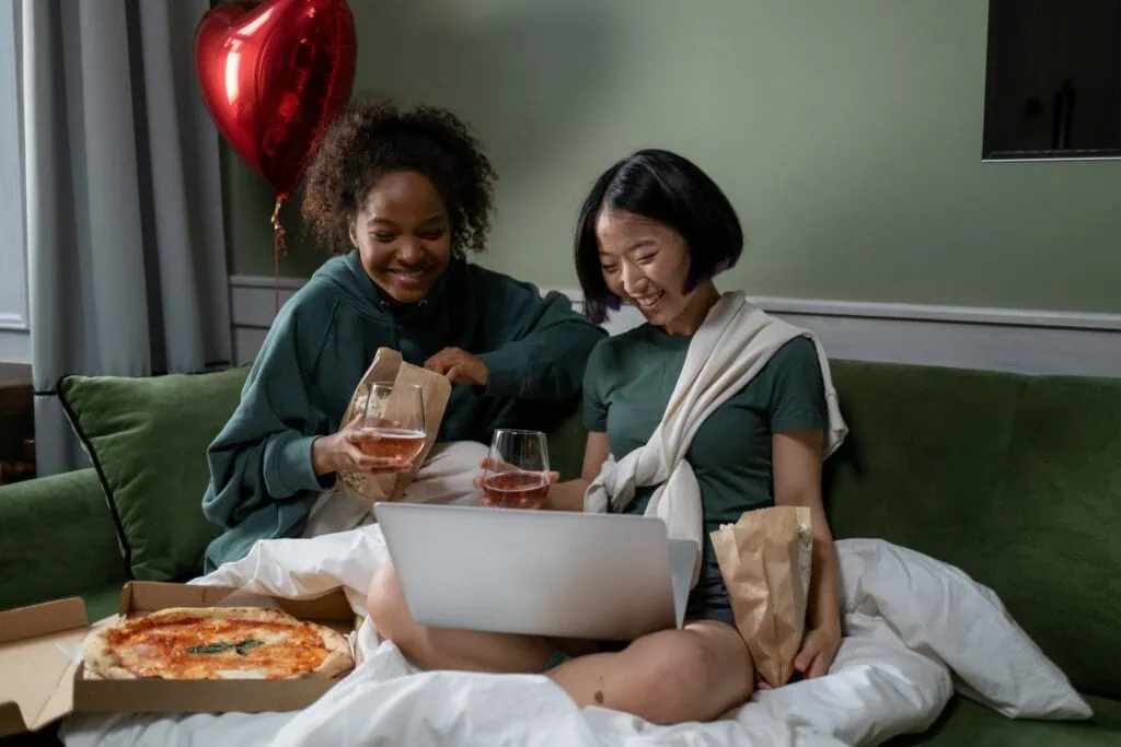 A couple watch something on a laptop while eating pizza and drinking wine on a couch.