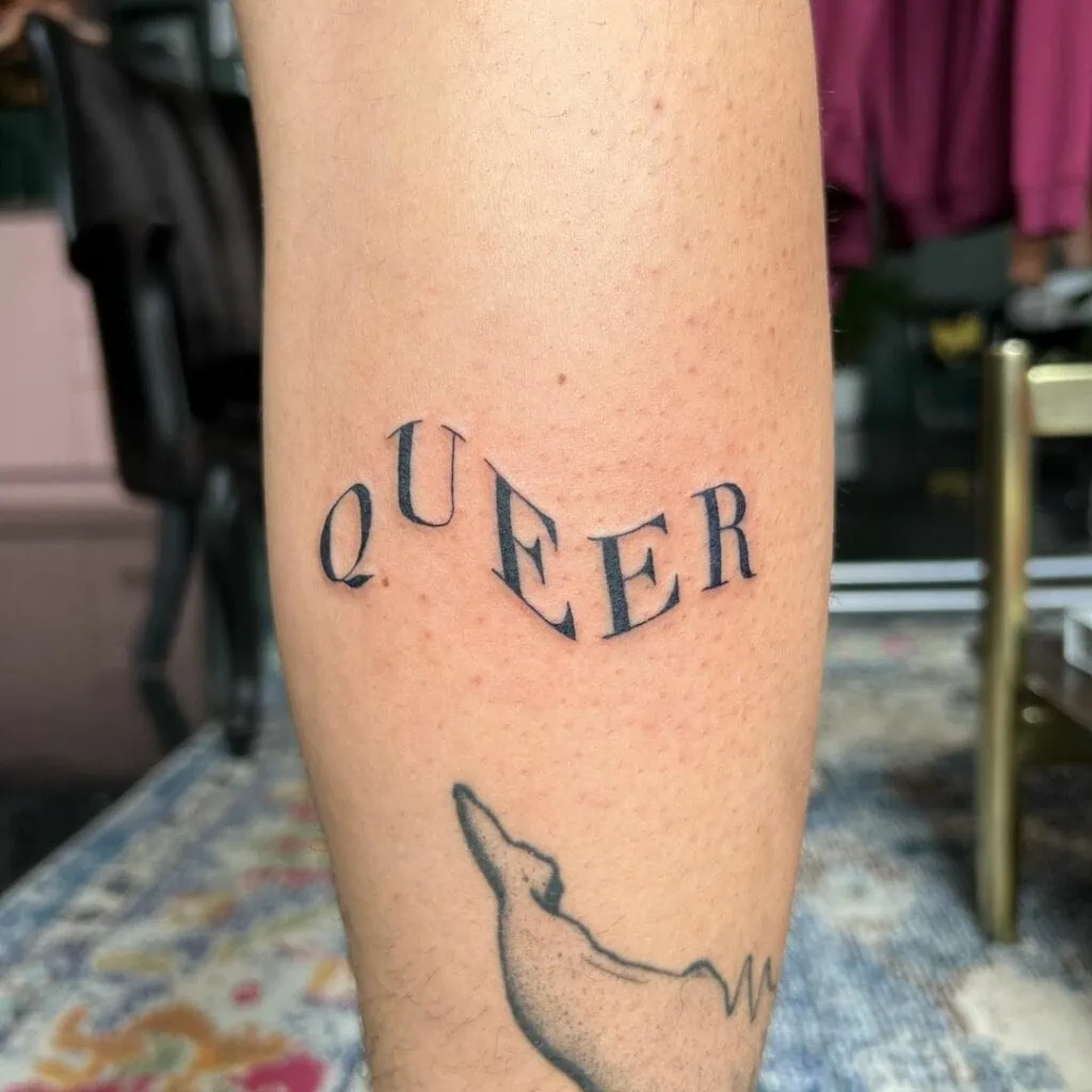 A simple LGBT tattoo reading “queer” in black ink
