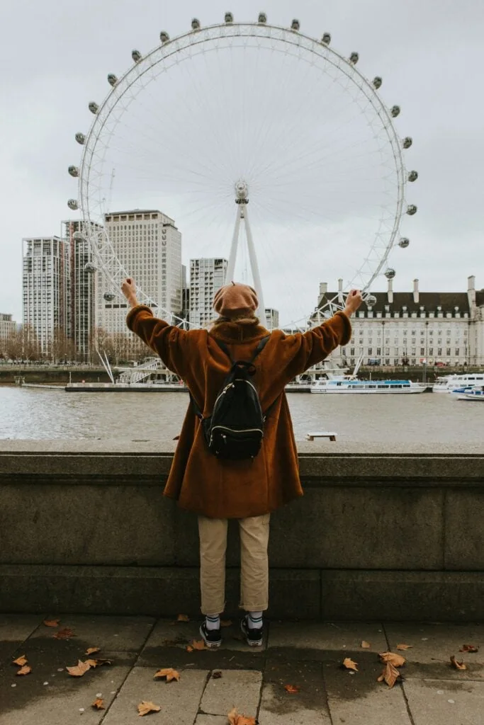A queer woman on the banks of the Thames with her arms outstretched towards the London Eye.
