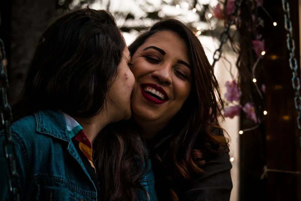 Two latina lesbians kissing and smiling with flowers and twinkle lights behind them.