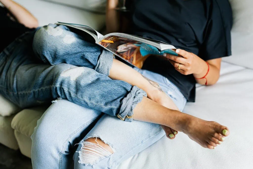 A queer couple sitting on a couch, both wearing jeans. One woman’s legs are resting on top of the other’s lap, while she reads a magazine.