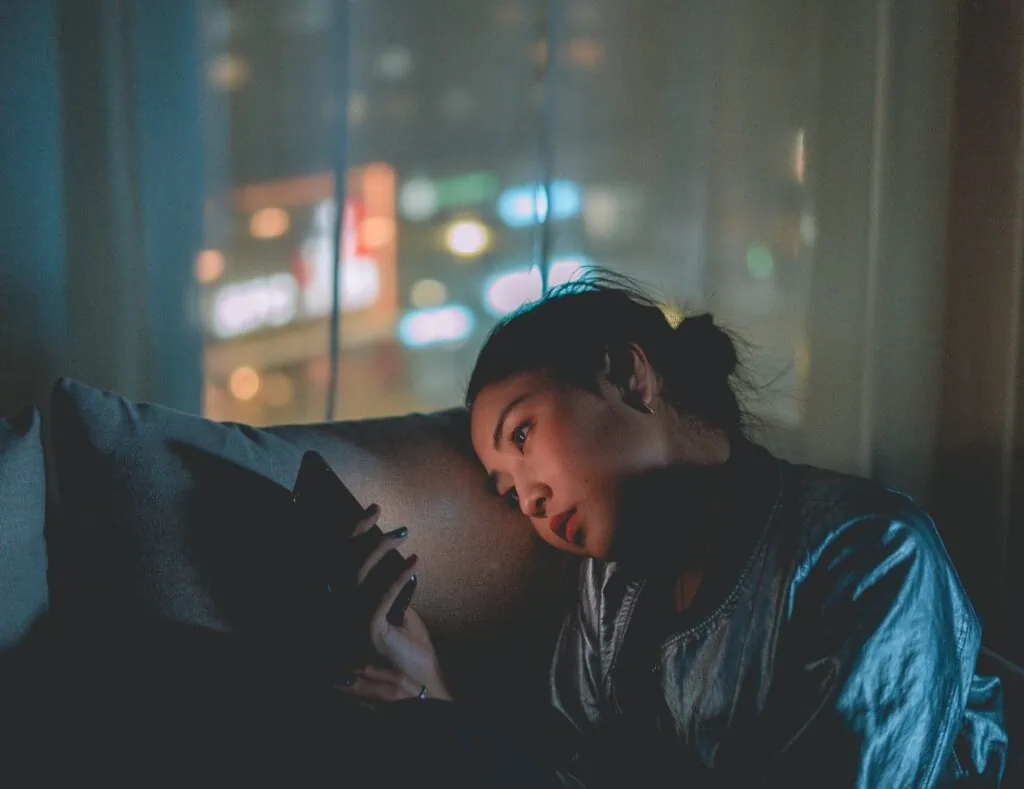 A queer Asian woman with a serious expression looking at her smart phone with a soft glow illuminating her face