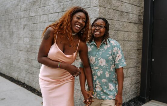 Two queer black women, one in a pink dress, the other in a teal and pink floral shirt, holding hands and laughing together