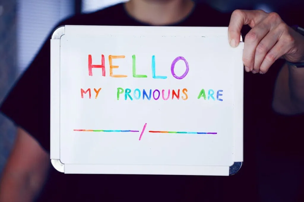 A person holds up a small whiteboard that reads “HELLO. MY PRONOUNS ARE,” followed by a black space with a slash dividing it. The text is written in rainbow colors.