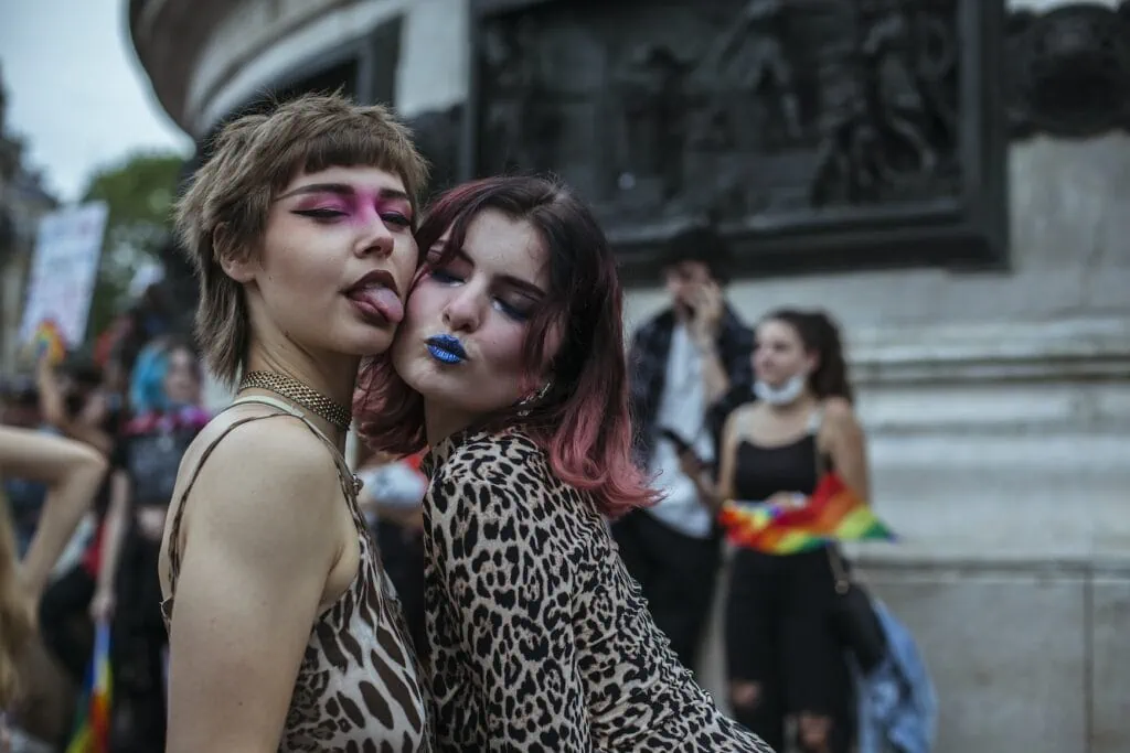 A couple wearing colorful makeup poses in front of the camera, with a crowd of behind them at a Pride march.
