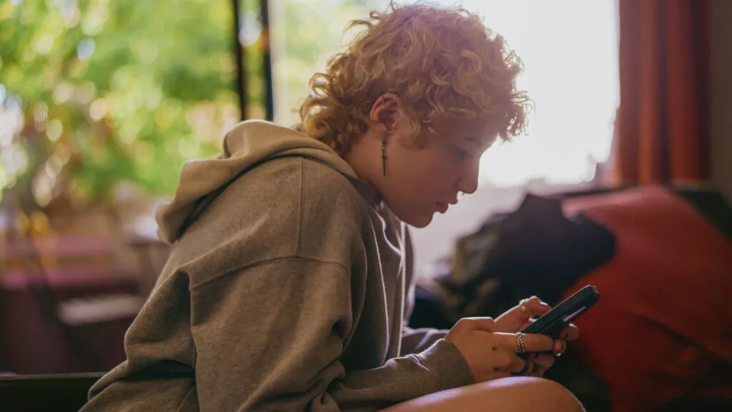 A non-binary person with short, blonde curly hair scrolls on their phone. They’re probably looking for new people to match with on the HER app.