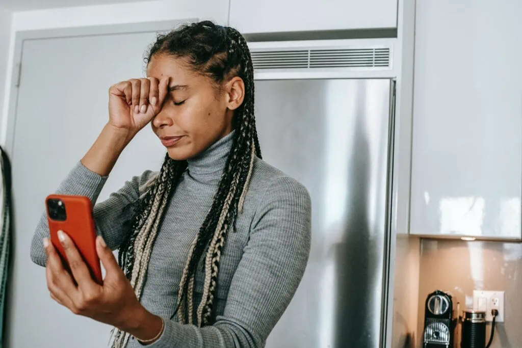 A black woman with braids looking at her phone in the kitchen with a frustrated look on her face.