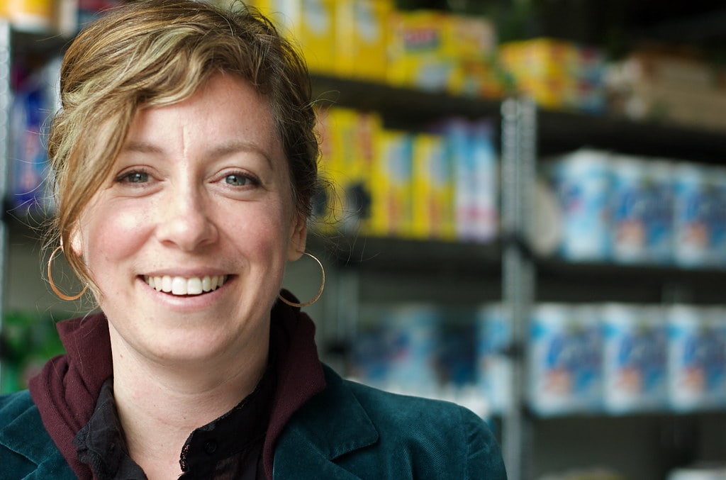 Une image de Thea Hillman, poète et activiste intersexe, dans une épicerie avec l'arrière-plan flou. Elle sourit et regarde l'appareil photo.
