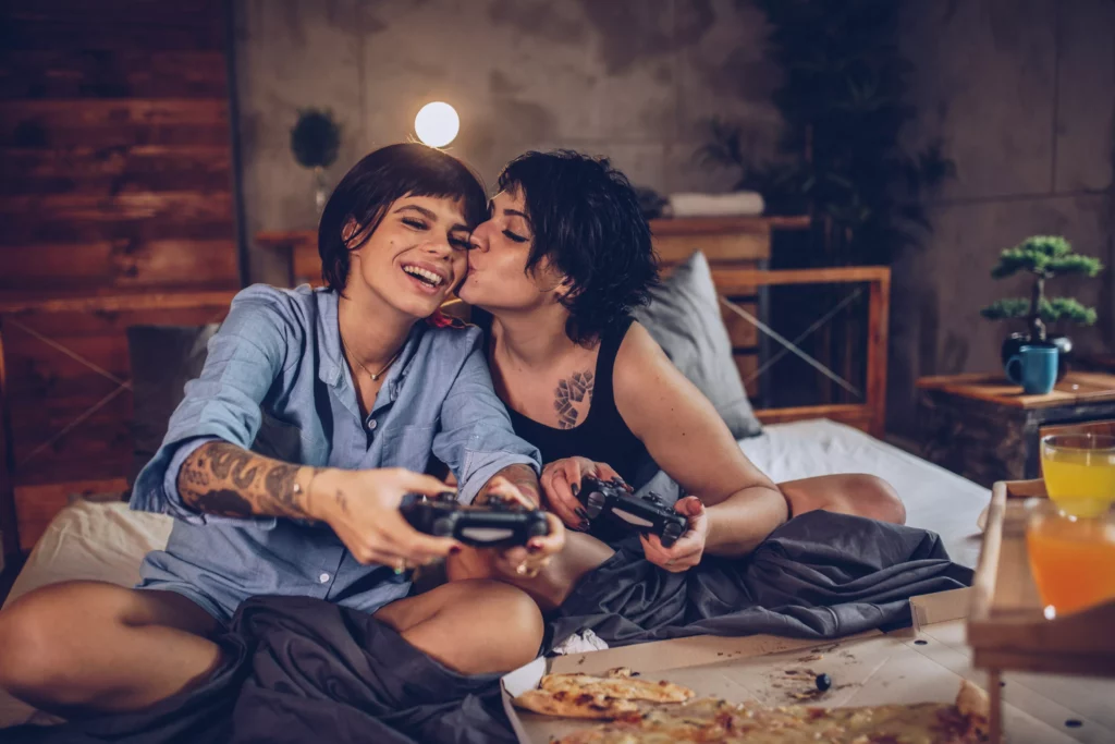 A couple of women playing video games in bed, with pizza and some drinks. Both have short hair and tattoos, one is kissing the other on the cheek.