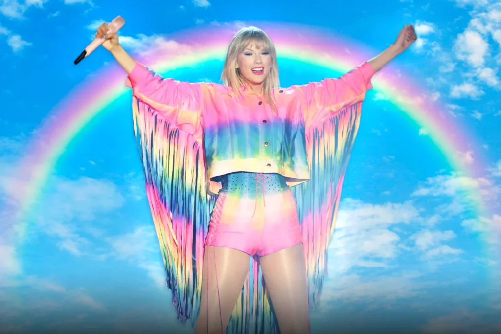 A picture of Taylor Swift in a pastel rainbow fringe costume with a rainbow behind her. Or is it Gaylor Swift now?