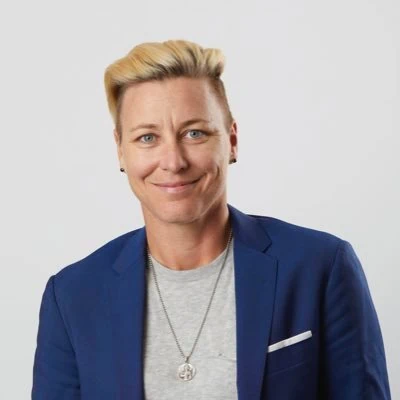 A headshot of Abby Wambach, who has short blonde hair and is wearing a navy blue blazer and a grey shirt.