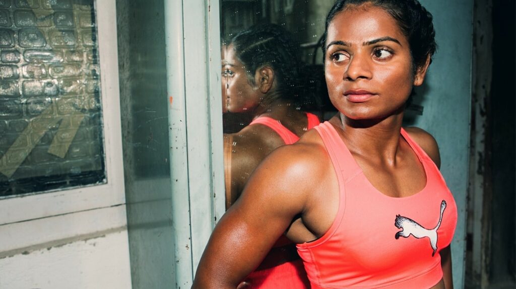 Dutee Chand, Indian professional sprinter and intersex woman, in a red Puma sports bra standing in front of a mirror relaxed, her muscles shining in the flash.