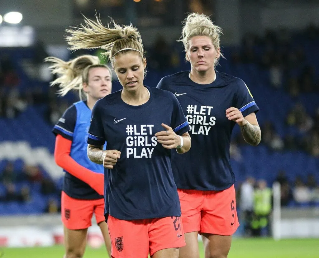 Rachel Daly at the front of two other teammates who are running in the middle of the soccer field, all wearing orange shorts and navy jerseys that read, #LetGirlsPlay.