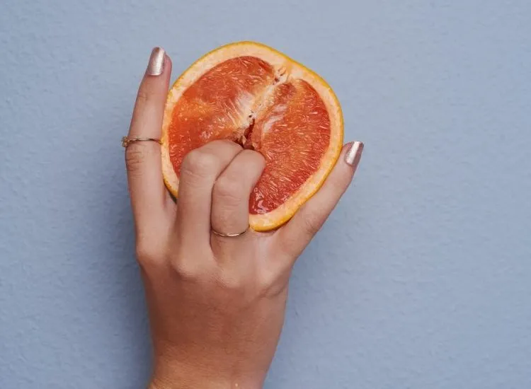 Fingers touching the middle of the half of a blood orange, over a pink background.