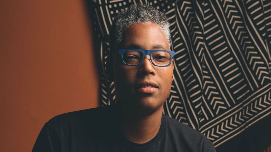An image of Sean Safia Wall in front of a black and brown tapestry. He is looking serious in blue glasses with salt and pepper hair, his brown skin glowing.