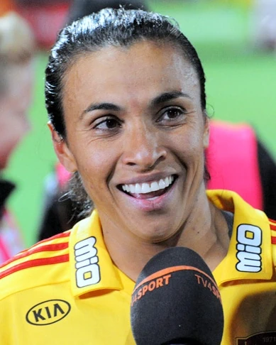 Marta wearing a yellow sports jersey, smiling as she speaks into a microphone after a game.