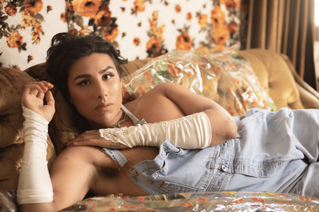 River Gallo posing on a couch in blue overalls and white gloves. They are laying down, looking relaxed with their arms slightly bent, looking at the camera.