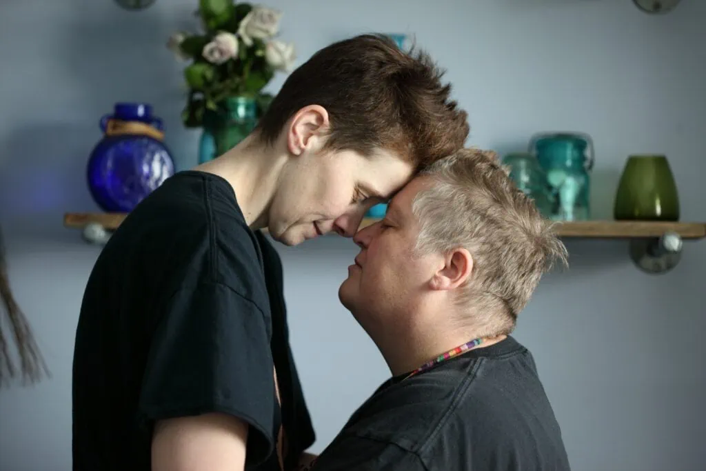 An older white butch lesbian introverted couple standing in front of a shelf with their foreheads pressed together.